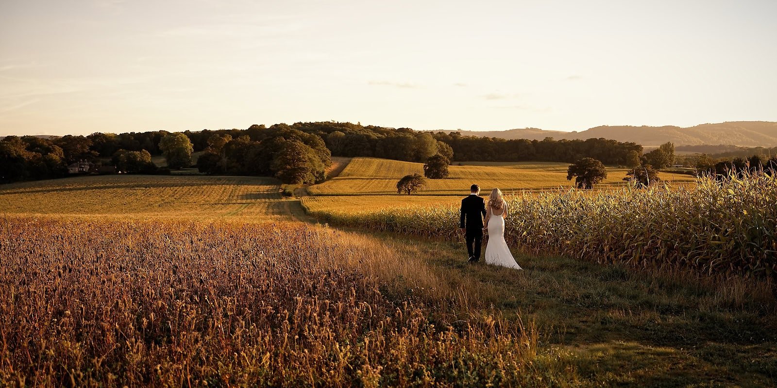 Bride and groom walking hand in hand into the cornfields behind Fitzleroi Barn at sunset