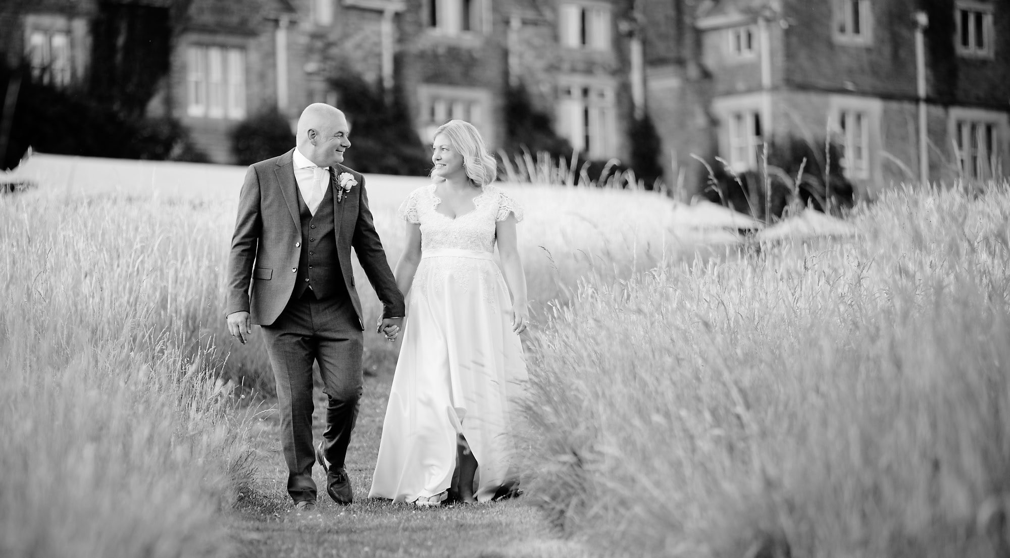 Black and white image of bride and groom walking through the long grass towards the lake at South Lodge Hotel, Horsham