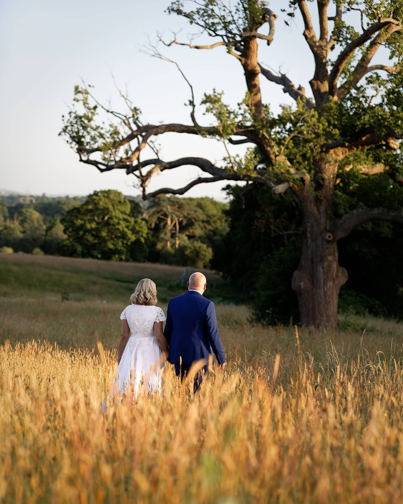 Bride and groom walking through the long grass with golden sunlight glowing on the tips at South Lodge, Horsham