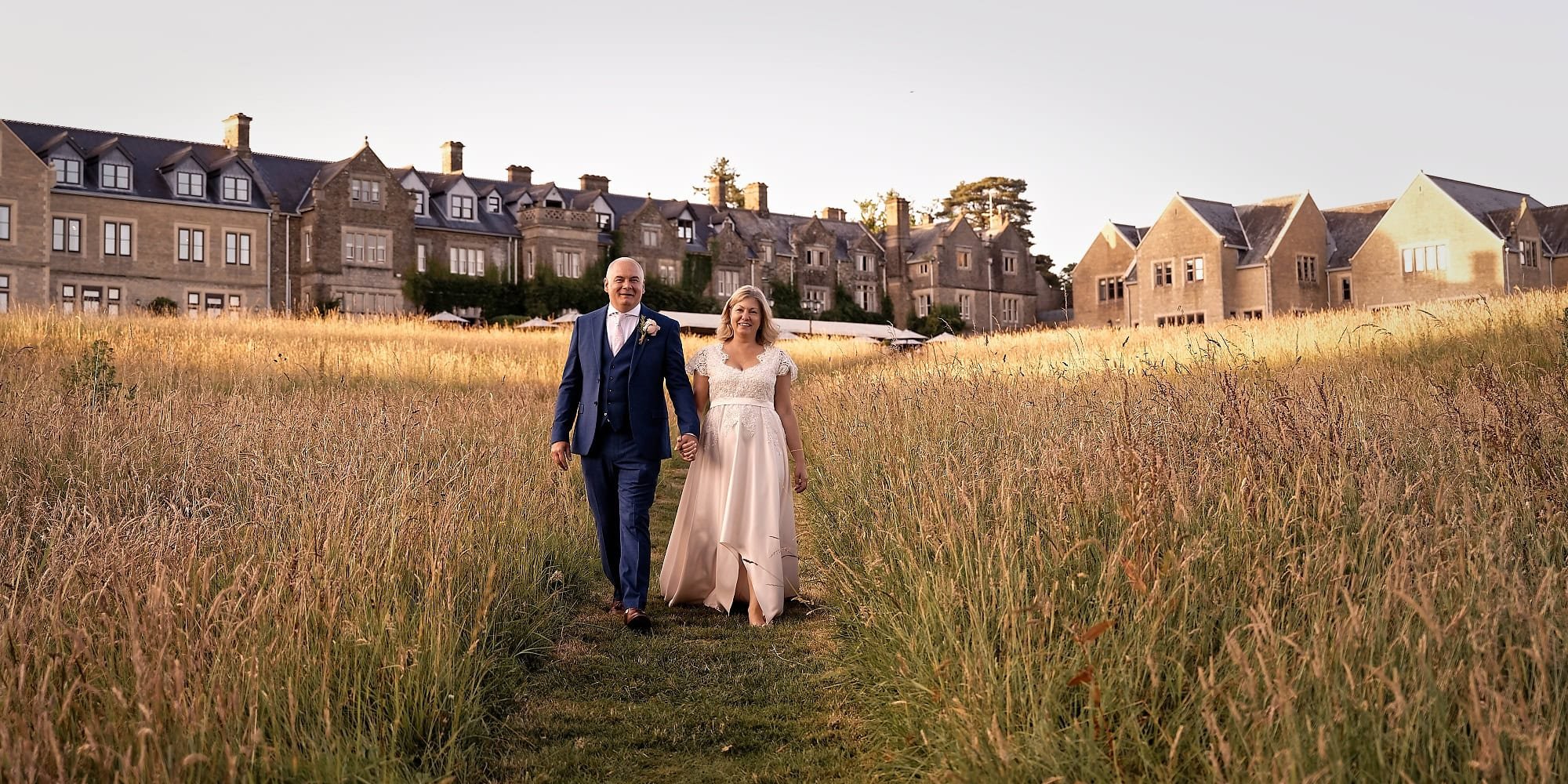 Bride and groom walking through the long grass on their wedding day with South Lodge in the background.