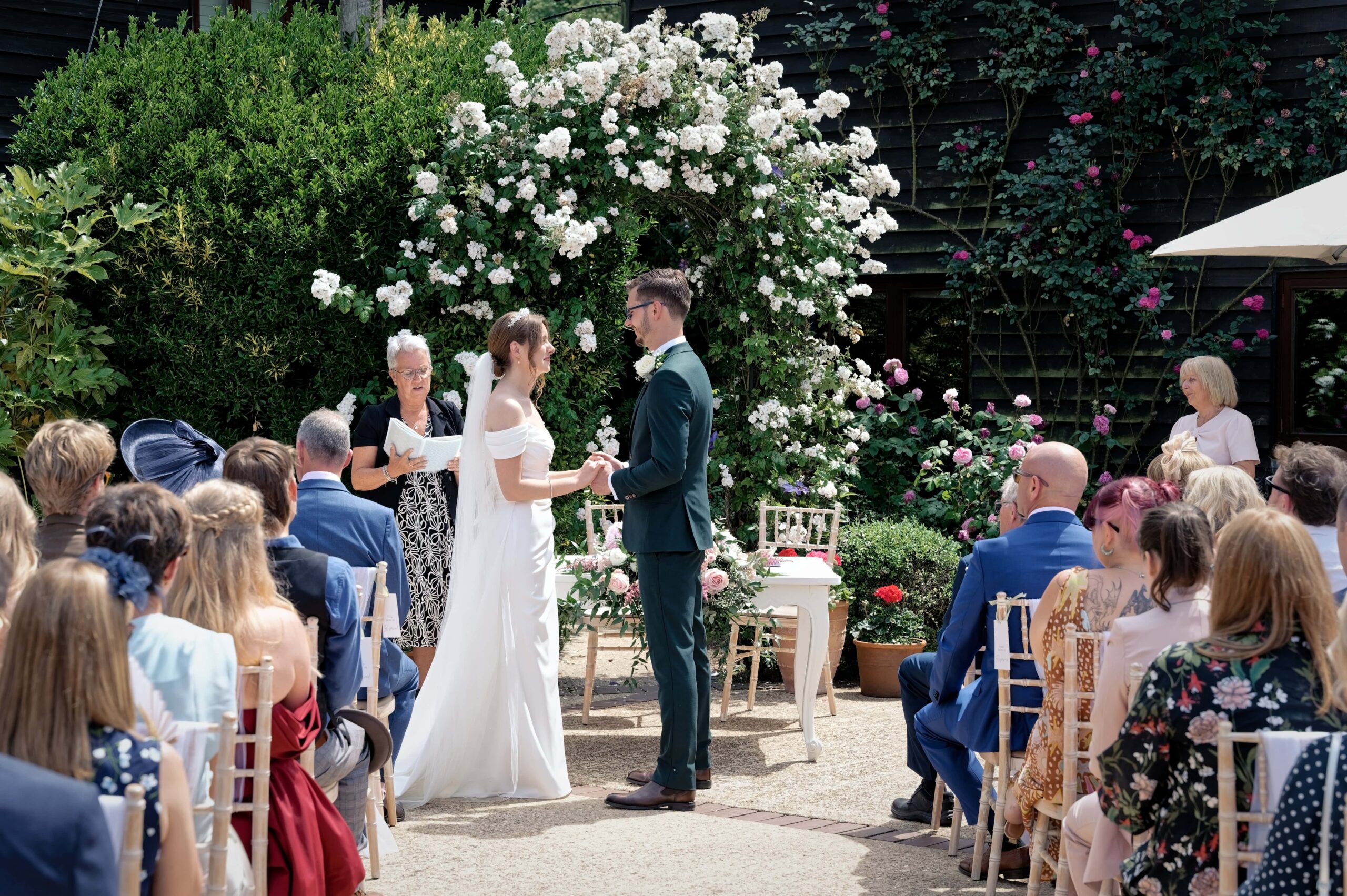 Fitzleroi Barn wedding photographer capturing an outdoor ceremony beneath a white rose archway
