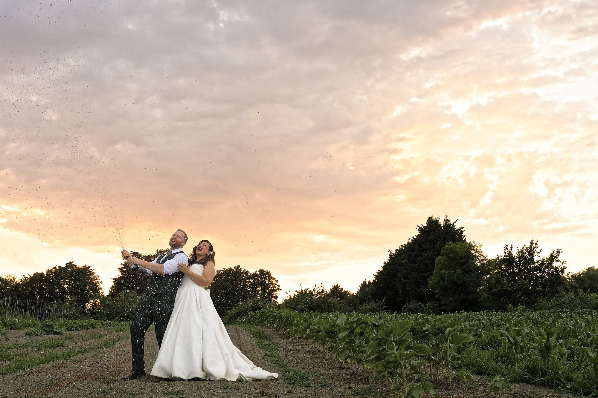Groom spraying champagne as the bride laughs beside him under a dramatic sunset sky
