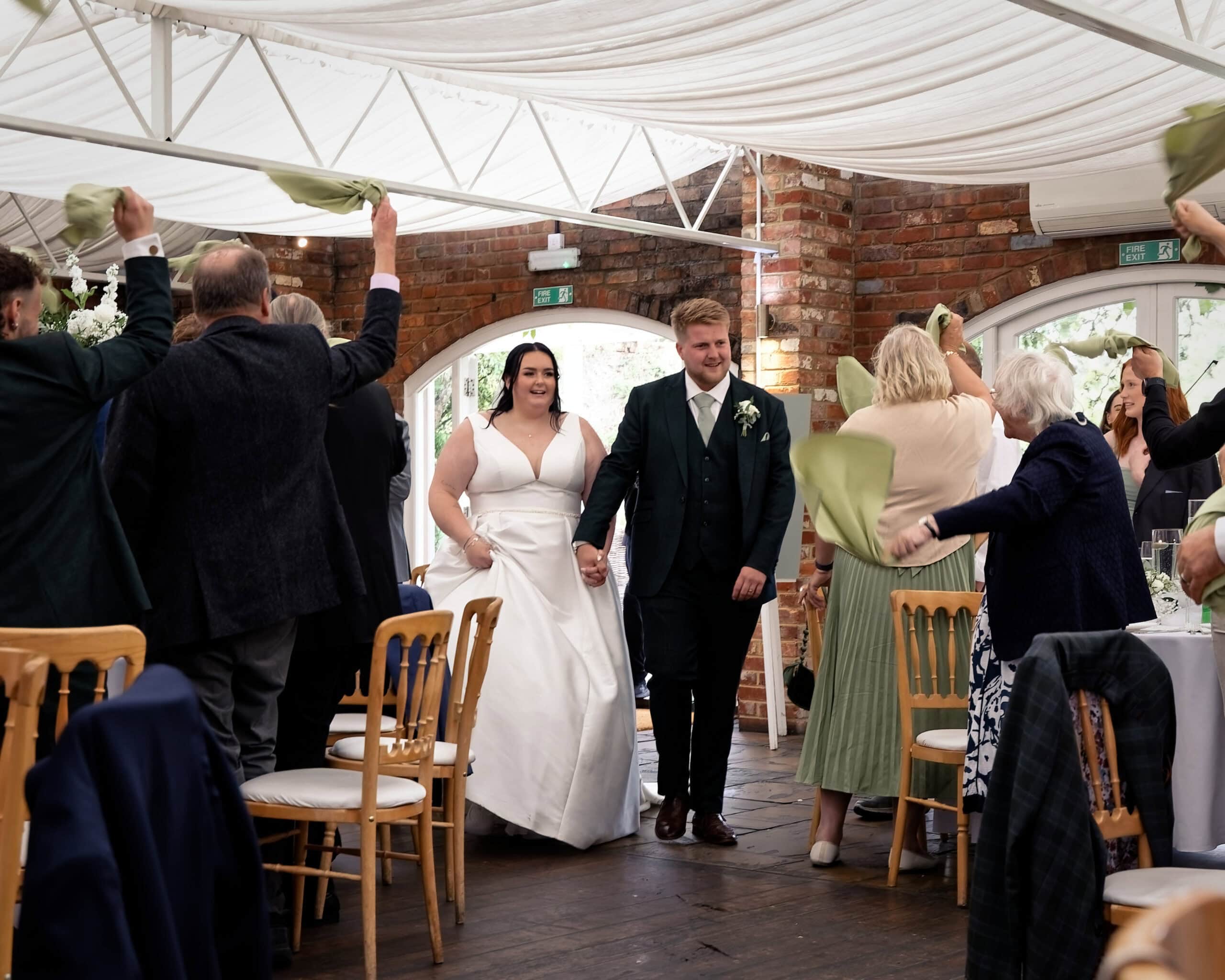 Meghan and Mark walking into the wedding breakfast room hand in hand as guests wave green napkins