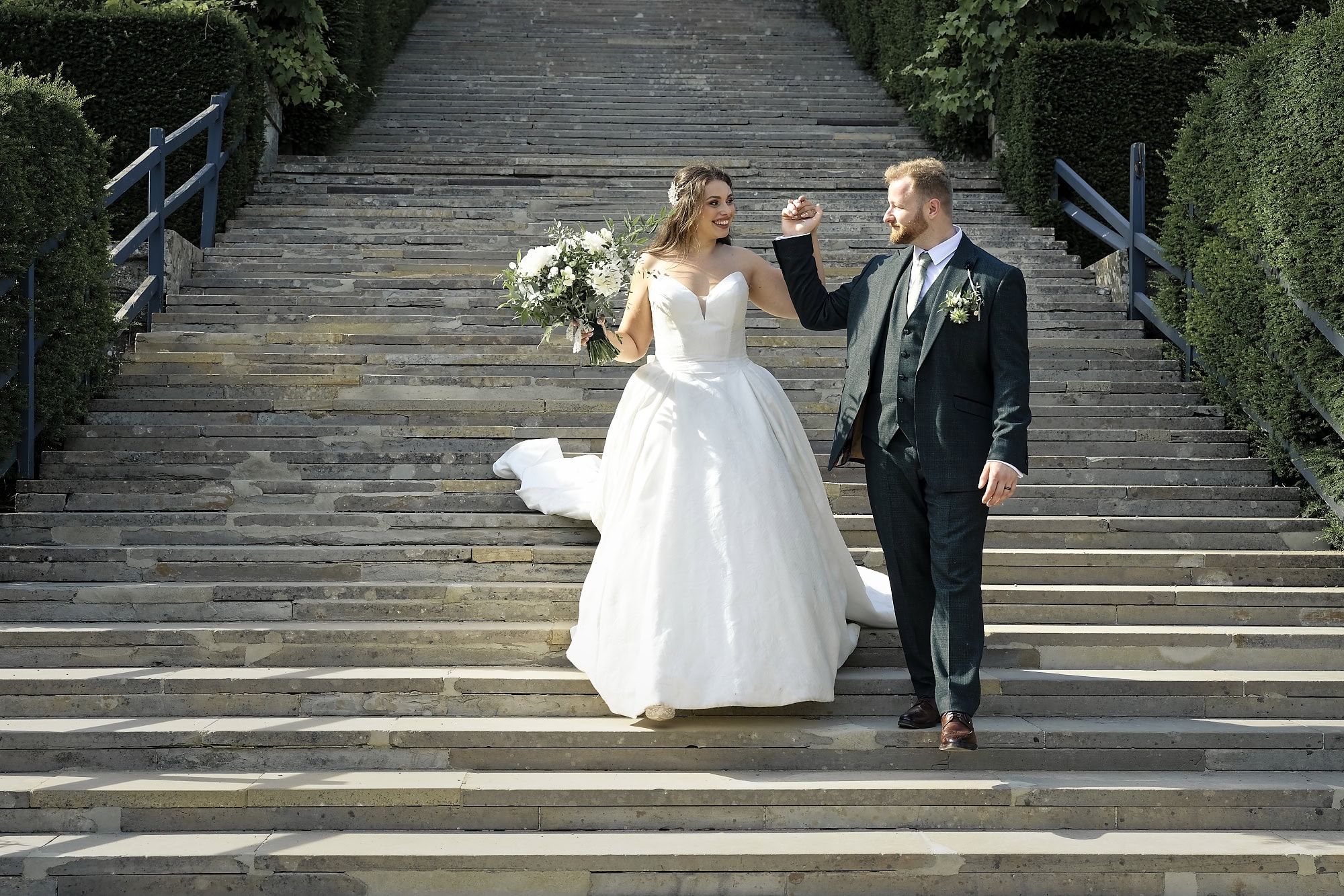 Bride and groom laughing together, his hand raised, walking down the grand steps at Port Lympne