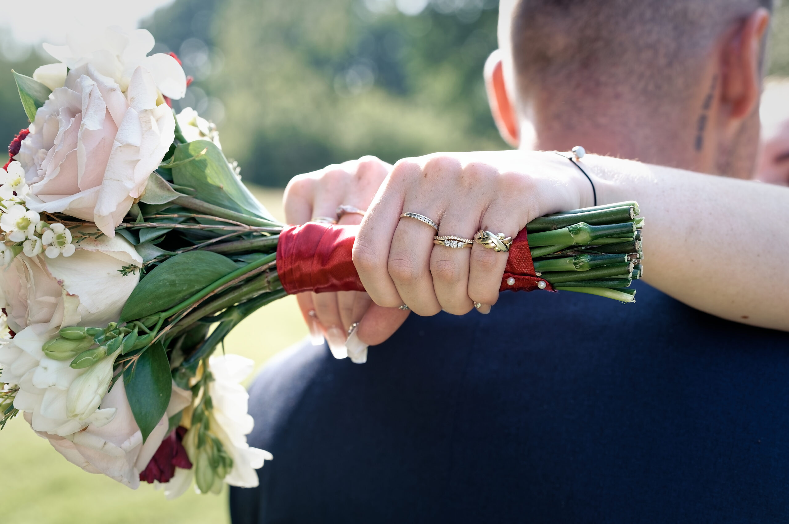 Bride's hand with wedding rings resting on the groom's shoulder, holding a blush and red bouquet at a Cottesmore Hotel wedding