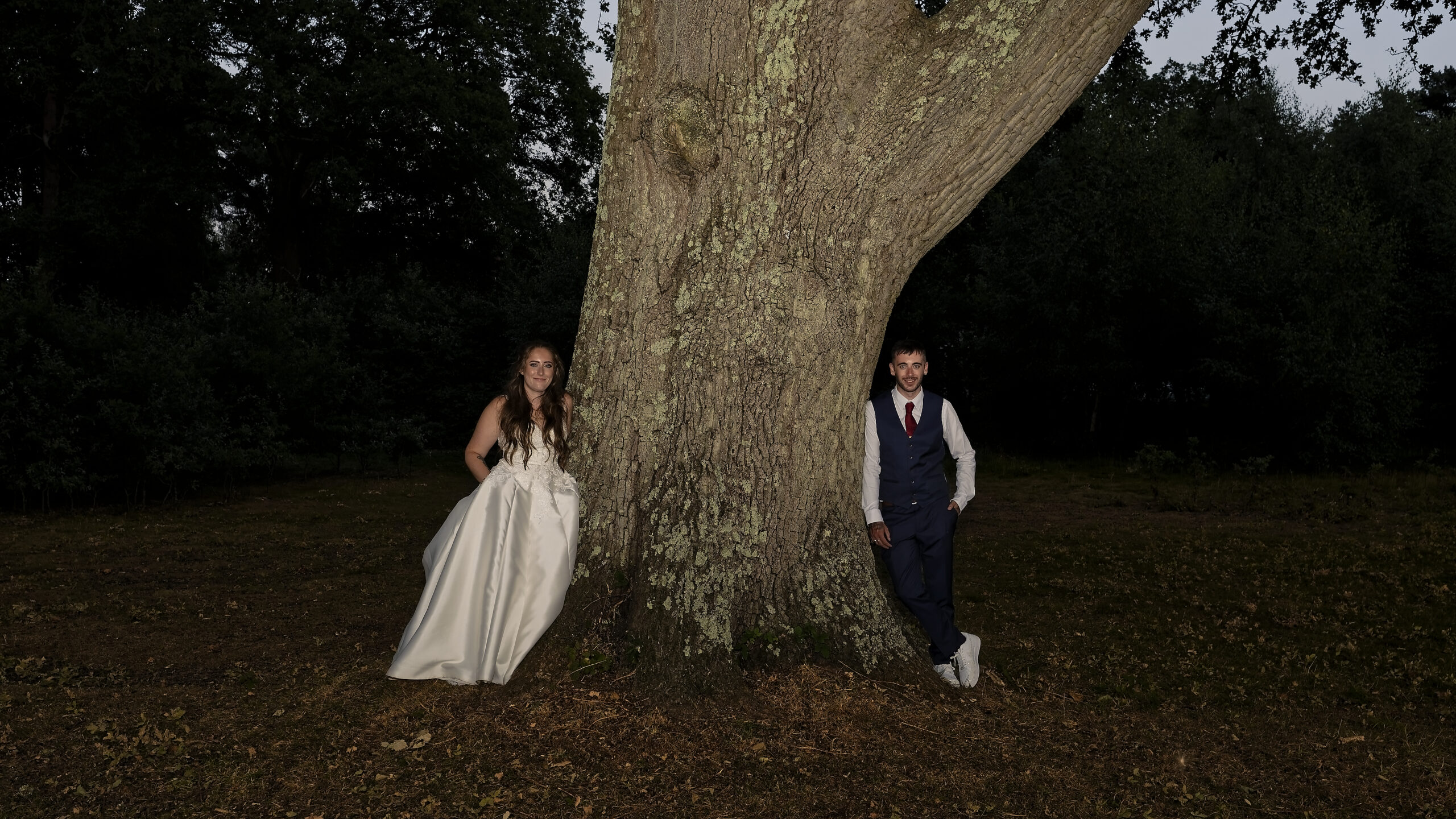 Bride and groom standing either side of a huge oak tree in the evening light at the Cottesmore Hotel