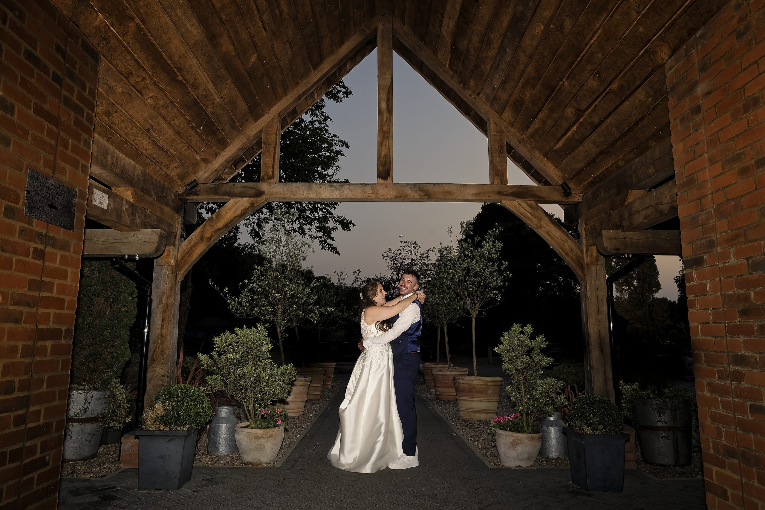 Bride and groom embracing under the oak beams of a timber barn entrance at dusk at the Cottesmore Hotel