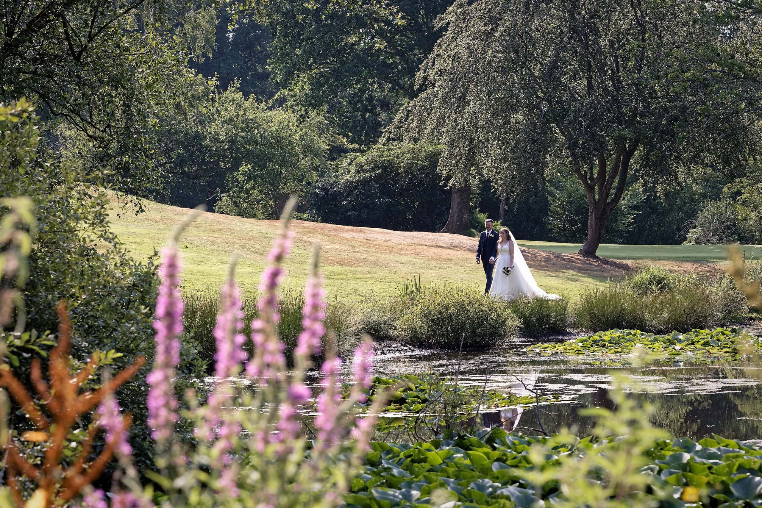 Bride and groom walking past pink flowers beside the lake, photographed by a Cottesmore Hotel wedding photographer