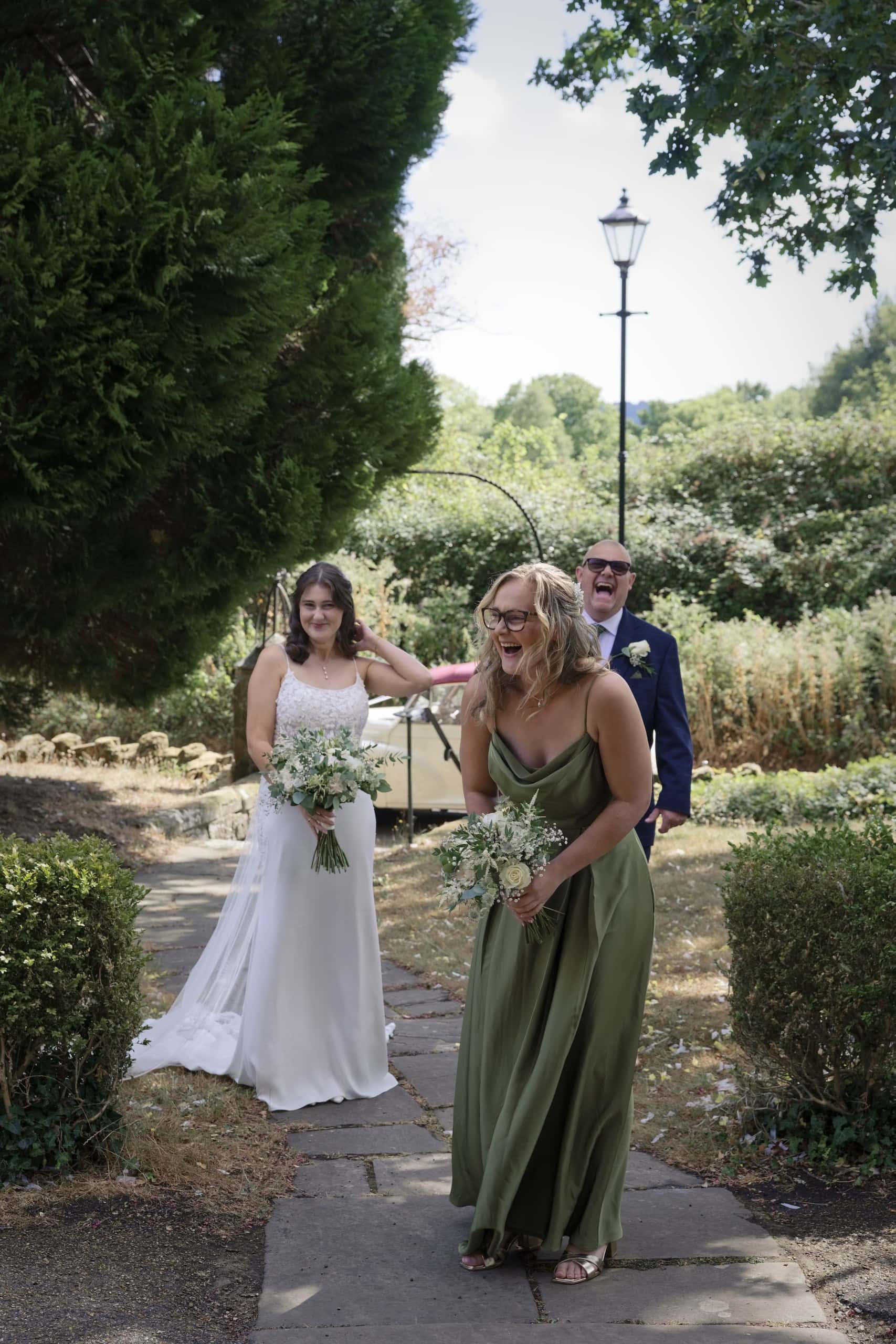Maid of honour in sage green laughing as the bride arrives at St Margaret of Antioch Church, Fernhurst