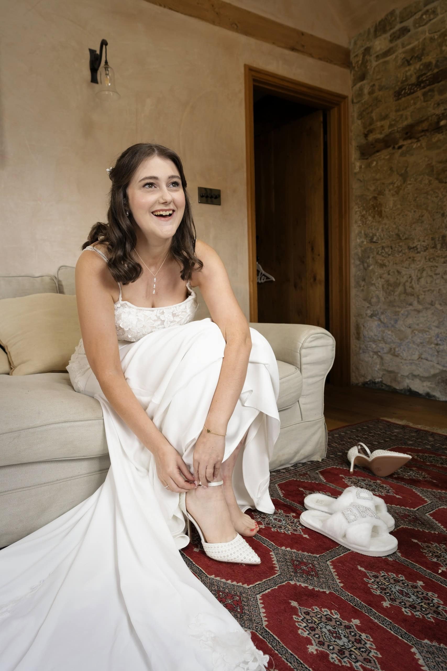 Bride laughing as she puts on her wedding shoes on a red patterned rug at Grittenham Barn
