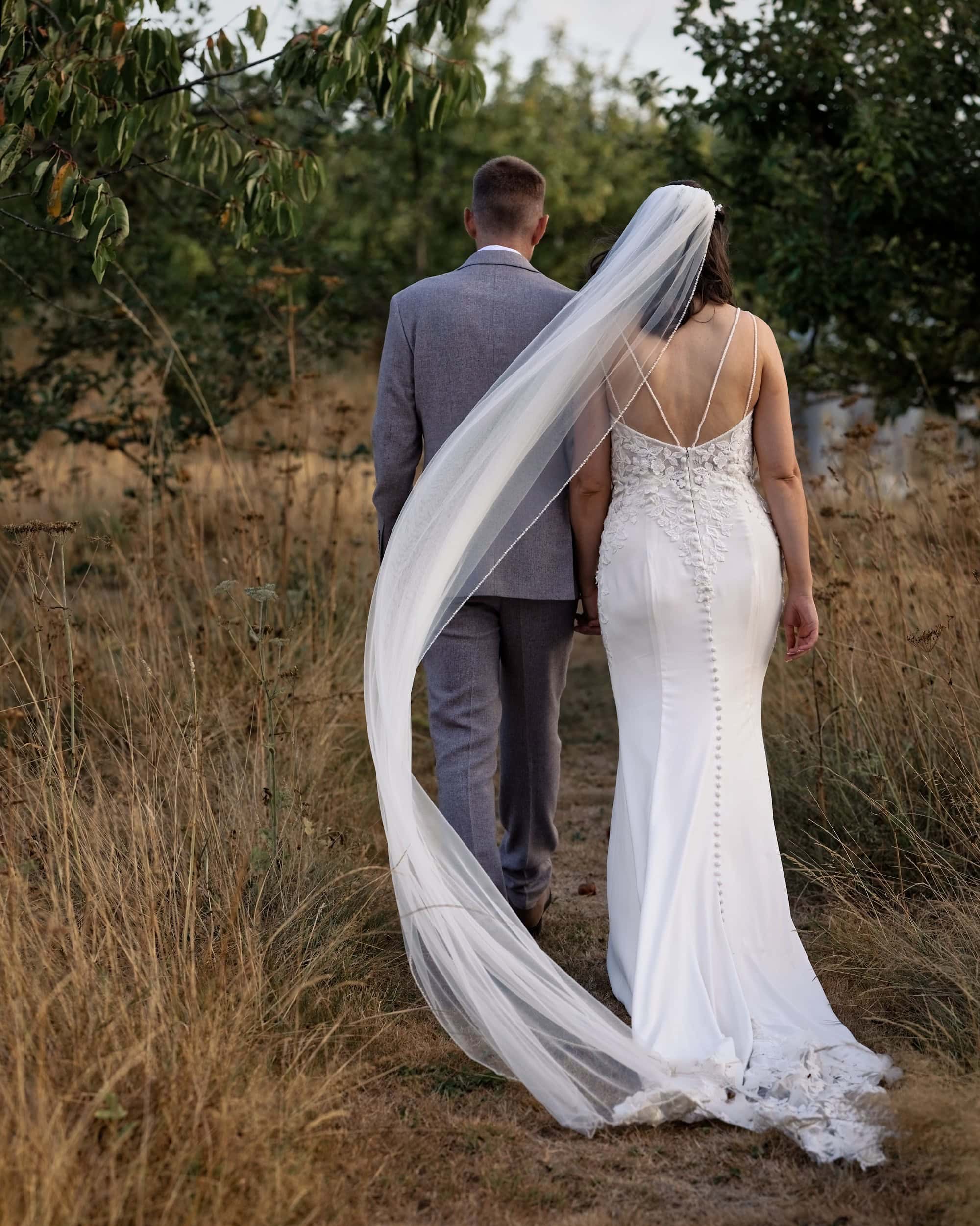 Long cathedral veil trailing behind the bride as the couple walk through the meadow
