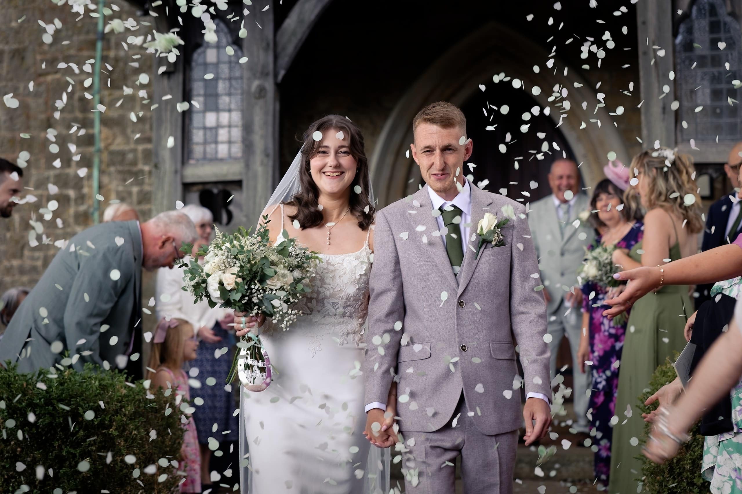 Bride and groom walking through a shower of white confetti outside St Margaret of Antioch Church