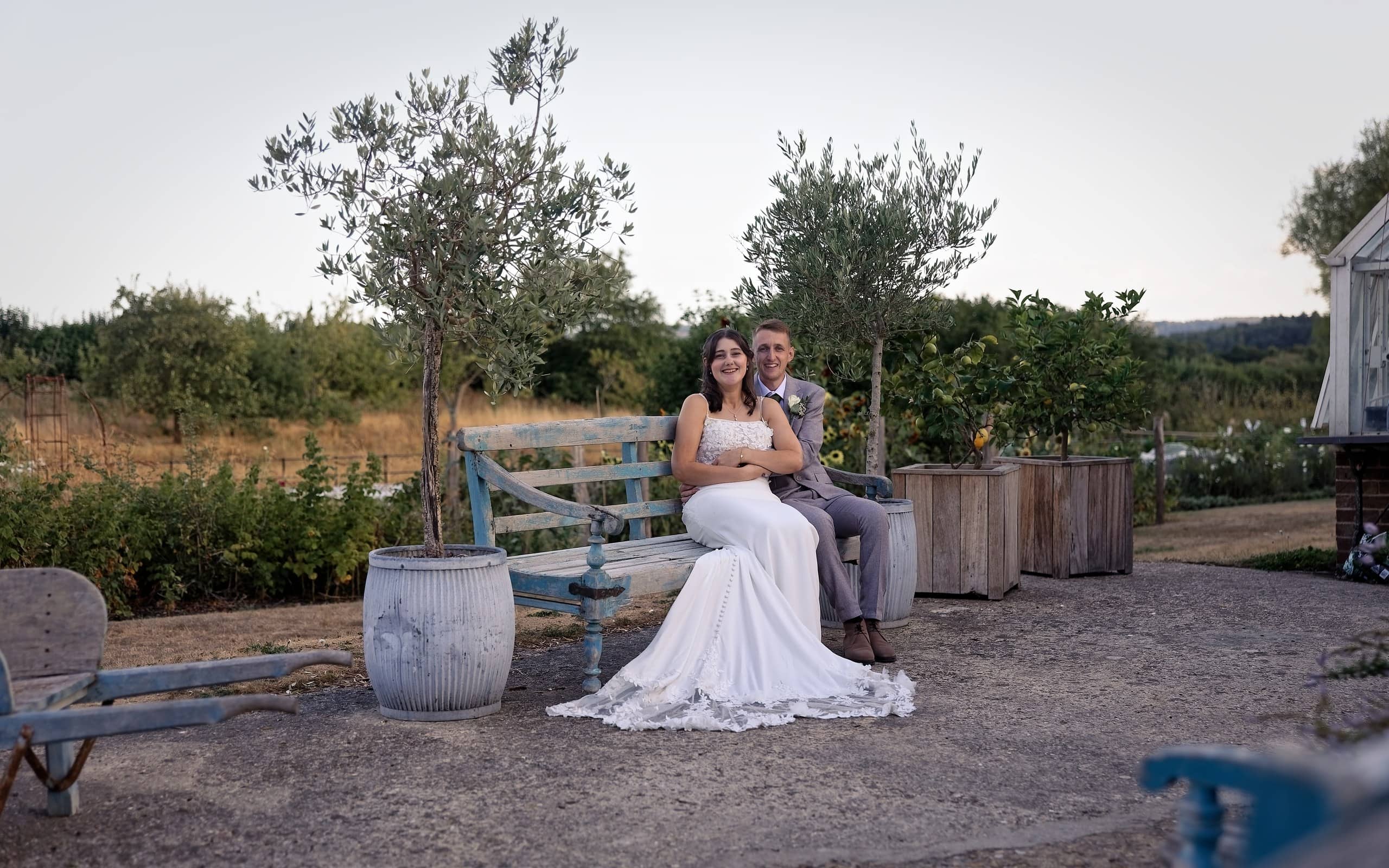 Newlyweds on a rustic bench among potted olive trees in the courtyard at Grittenham Barn