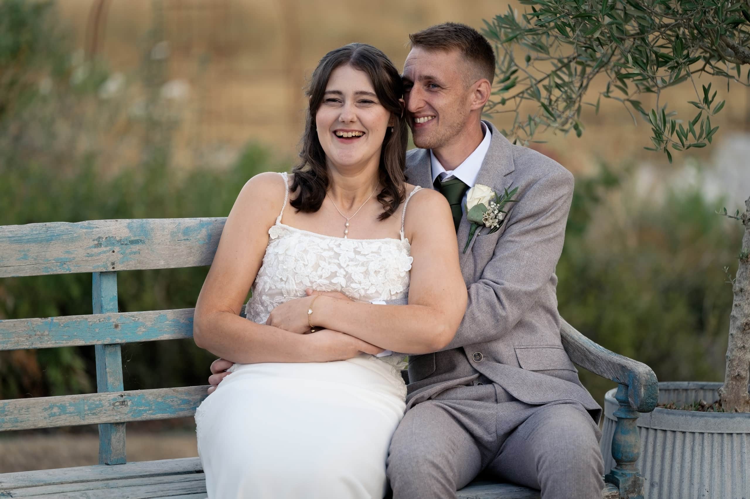 Bride and groom laughing together on a weathered blue bench at Grittenham Barn