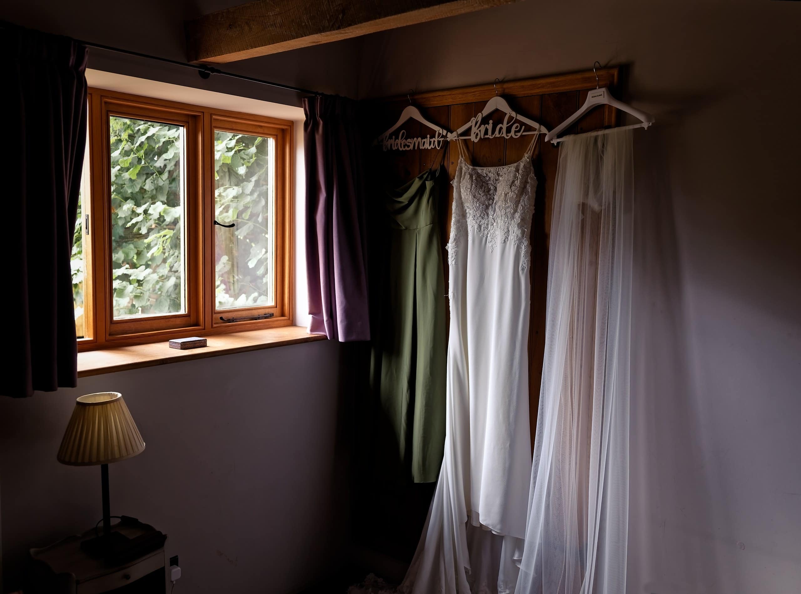 Wedding dress and sage green bridesmaid dress hanging by the window at Grittenham Barn