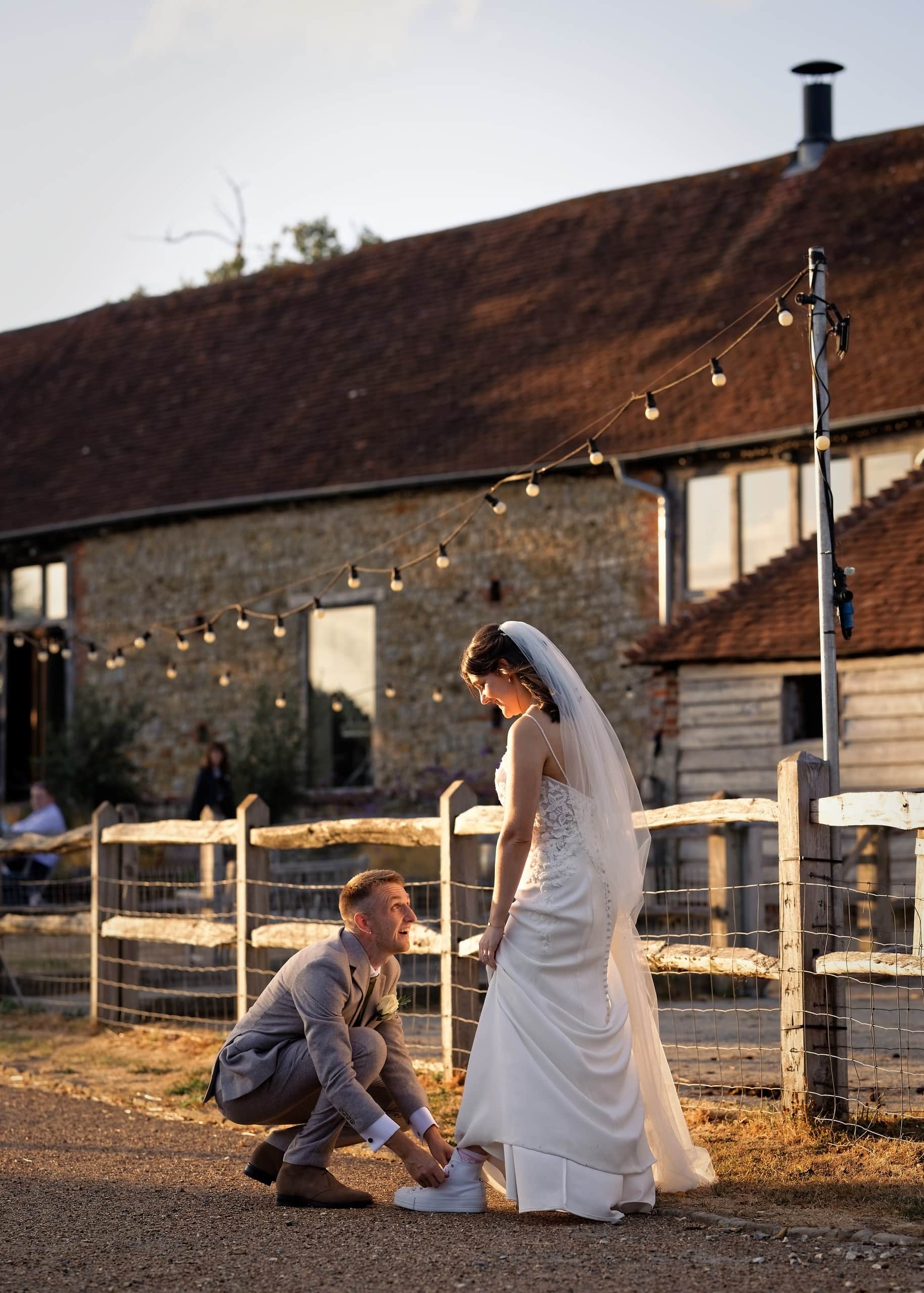 Groom kneeling to help the bride with her shoes outside the barn under festoon lights