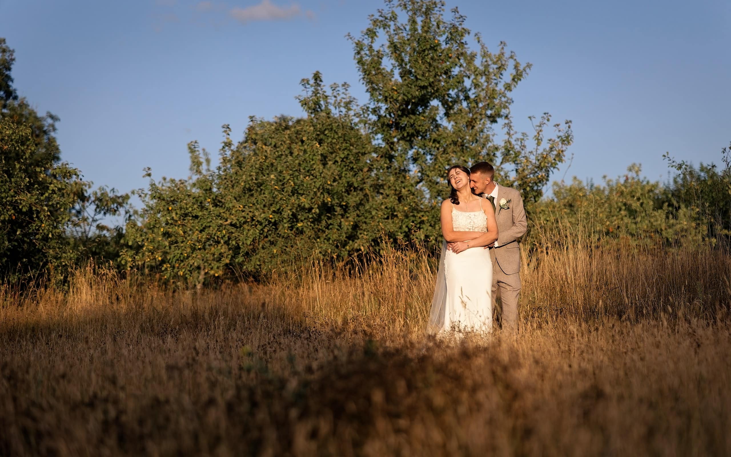 Bride and groom embracing in warm sunset light in the long grass at Grittenham Barn